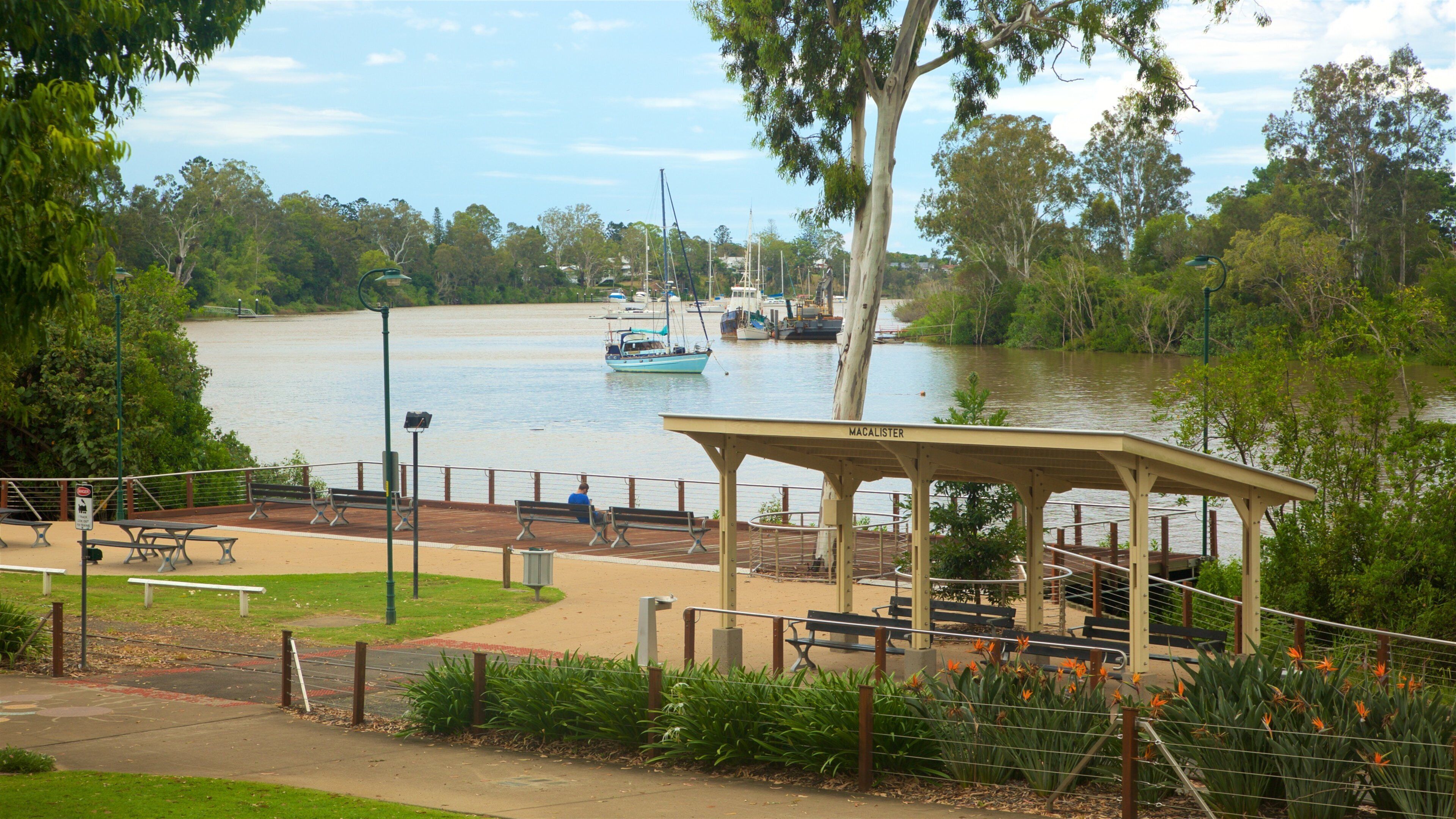 Un parque junto al río con bancos, un quiosco y barcos en el agua.