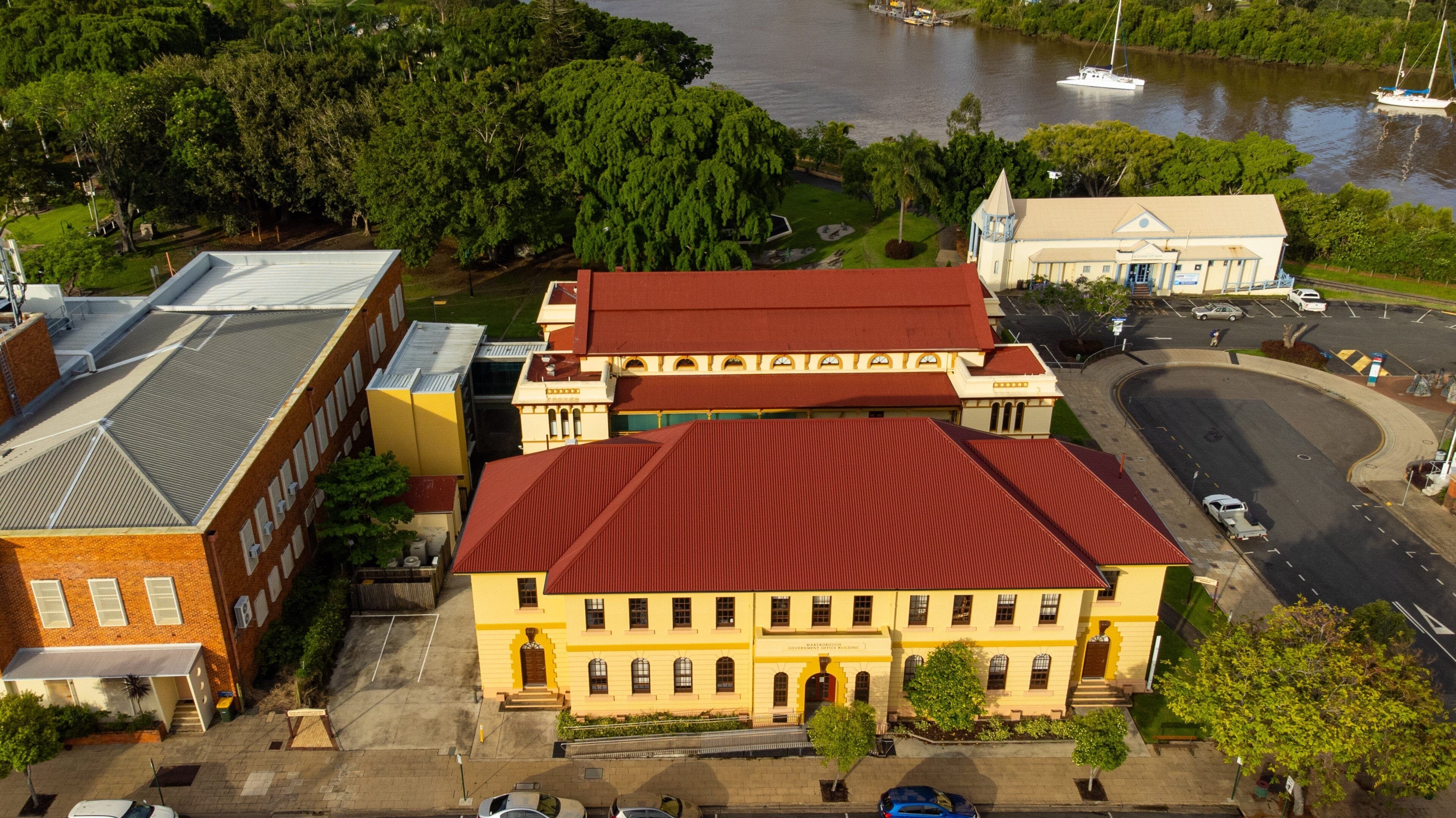 A large yellow building with a red roof, surrounded by trees and a river.