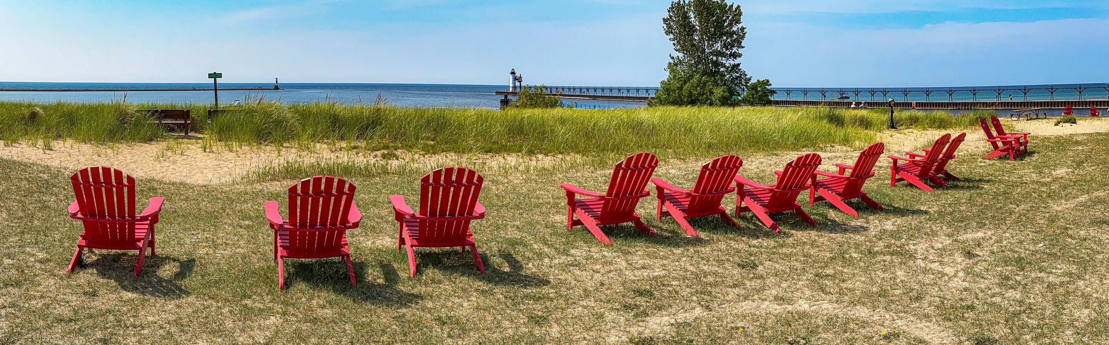 A row of red Adirondack chairs on a grassy beach with a lighthouse and pier in the background.