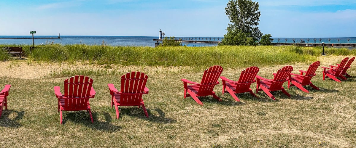 A row of red Adirondack chairs on a grassy beach with a lighthouse and pier in the background.
