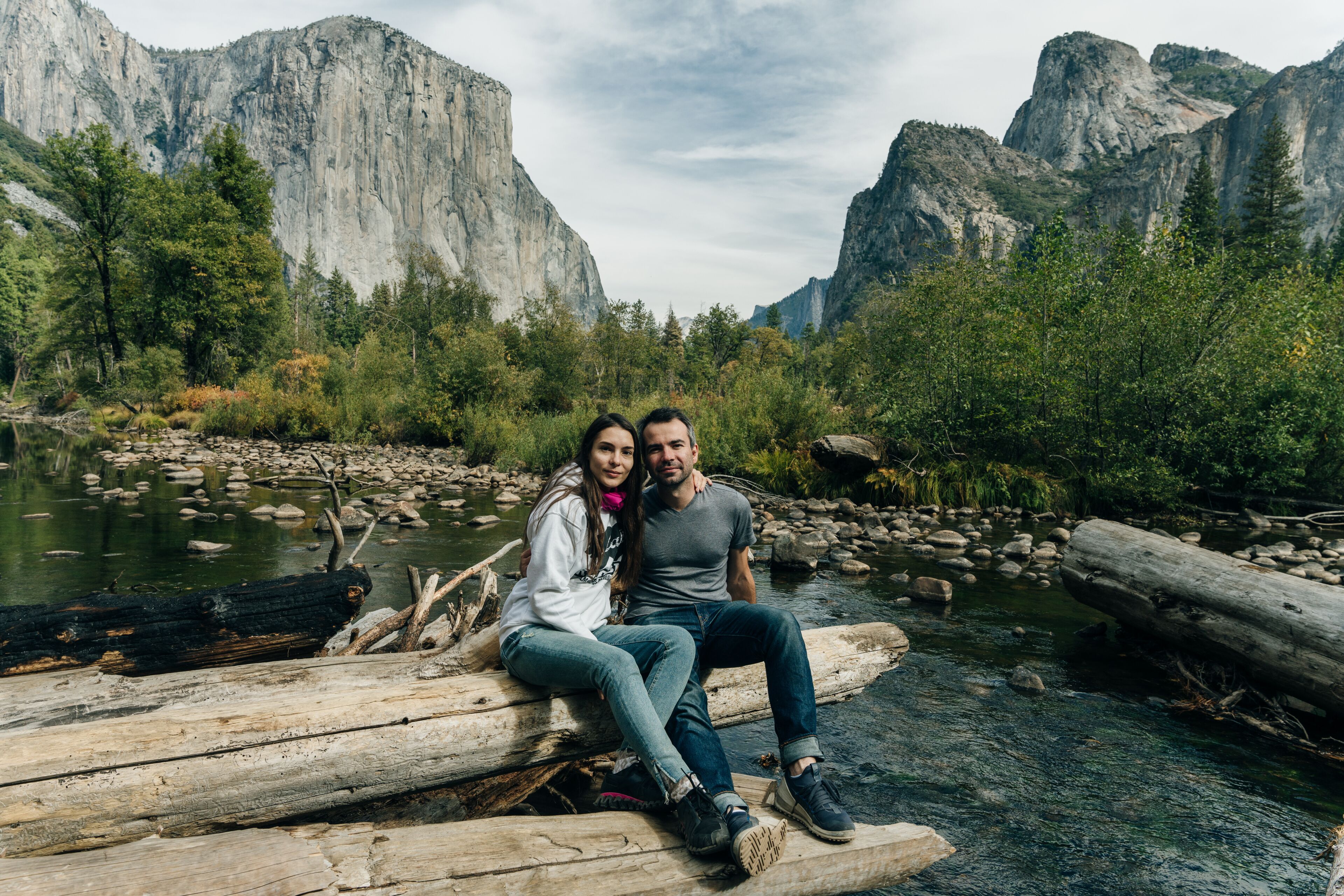 A couple sitting on a log by a river with mountains in the background.