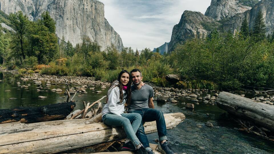 A couple sitting on a log by a river with mountains in the background.