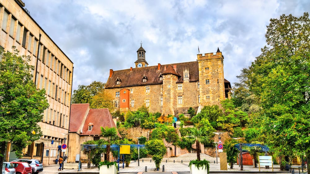 A historic building with a clock tower, surrounded by lush greenery and a paved square.