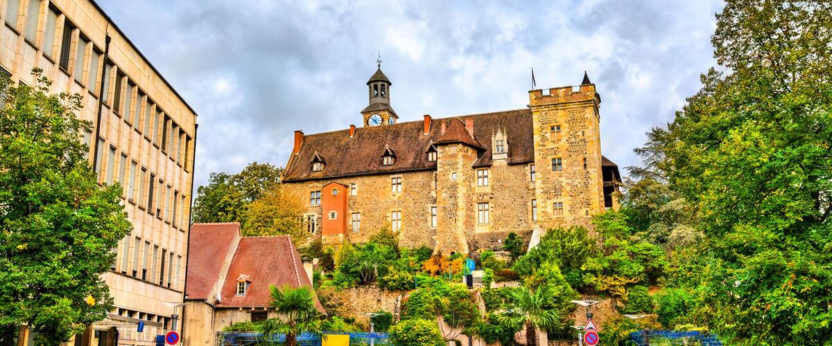 A historic building with a clock tower, surrounded by lush greenery and a paved square.