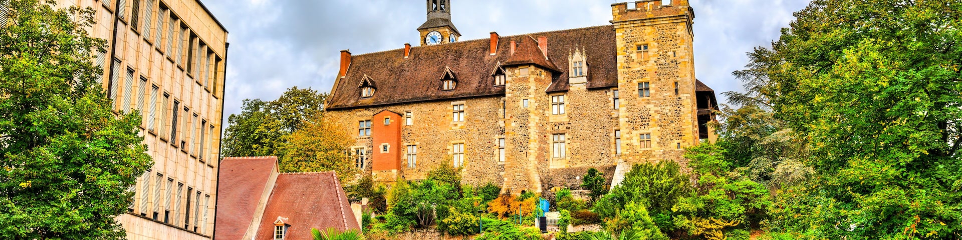 A historic building with a clock tower, surrounded by lush greenery and a paved square.