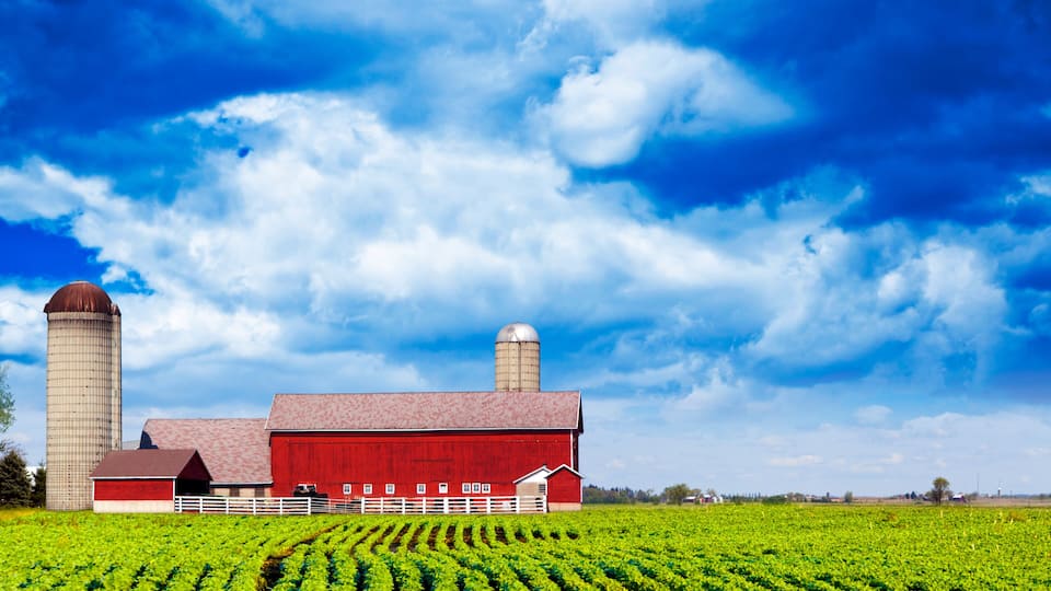 A red barn with a silo, surrounded by green fields under a blue sky with clouds.