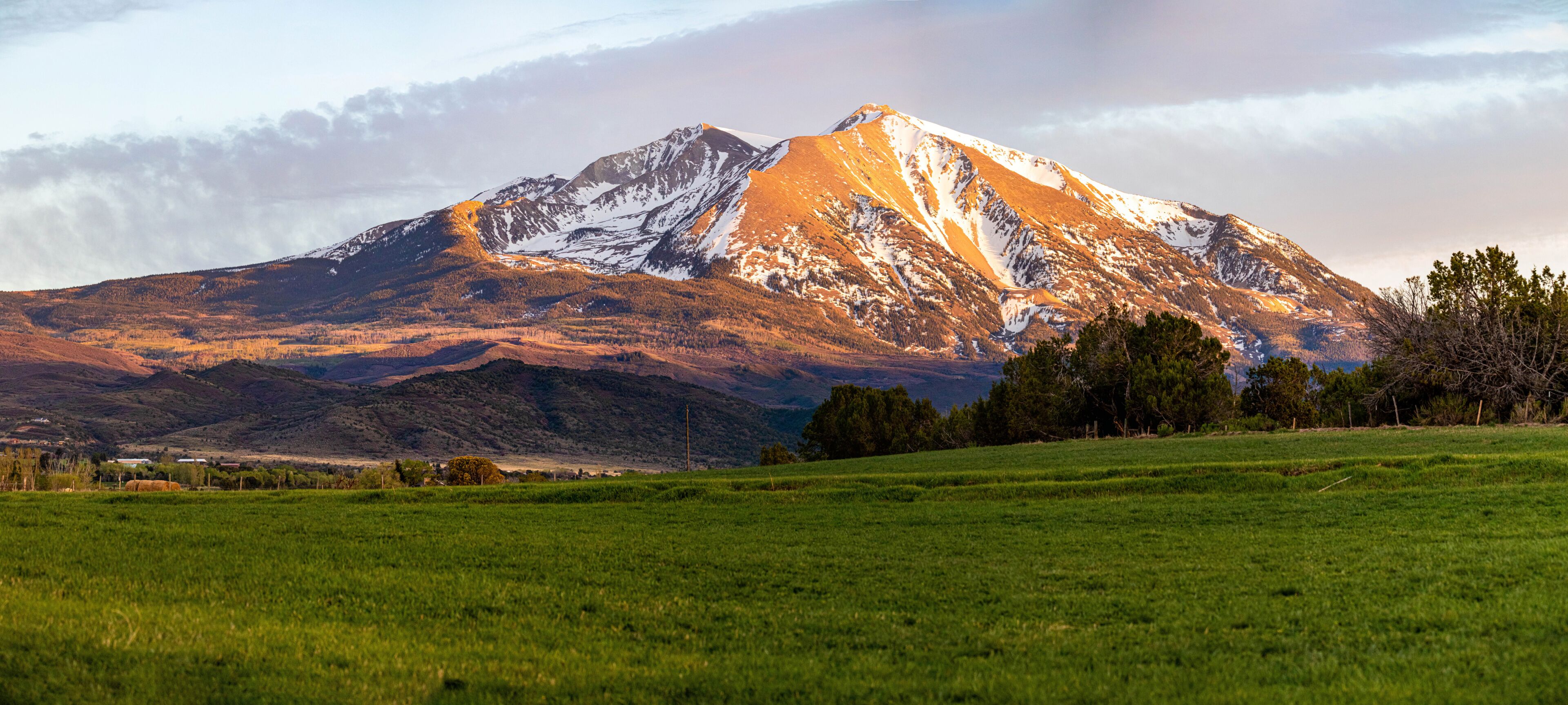 A snow-capped mountain with a green foreground field and distant buildings.