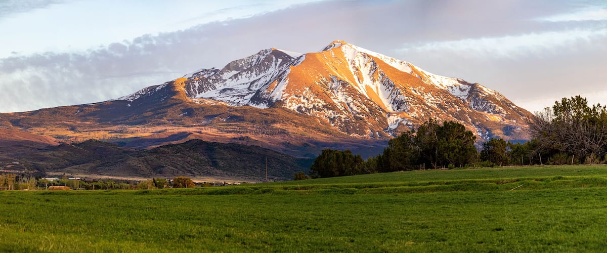 A snow-capped mountain with a green foreground field and distant buildings.