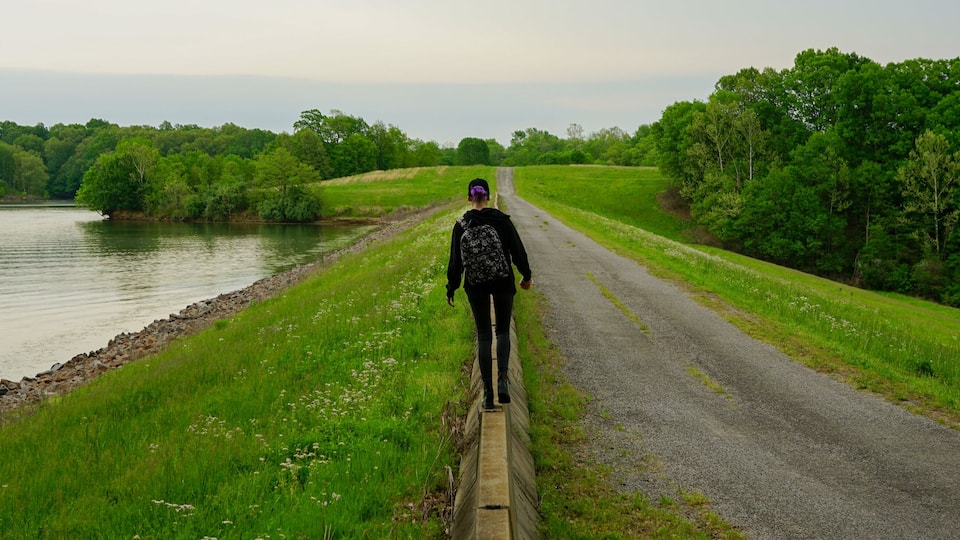 A person walking along a gravel path beside a river, surrounded by lush greenery and trees.