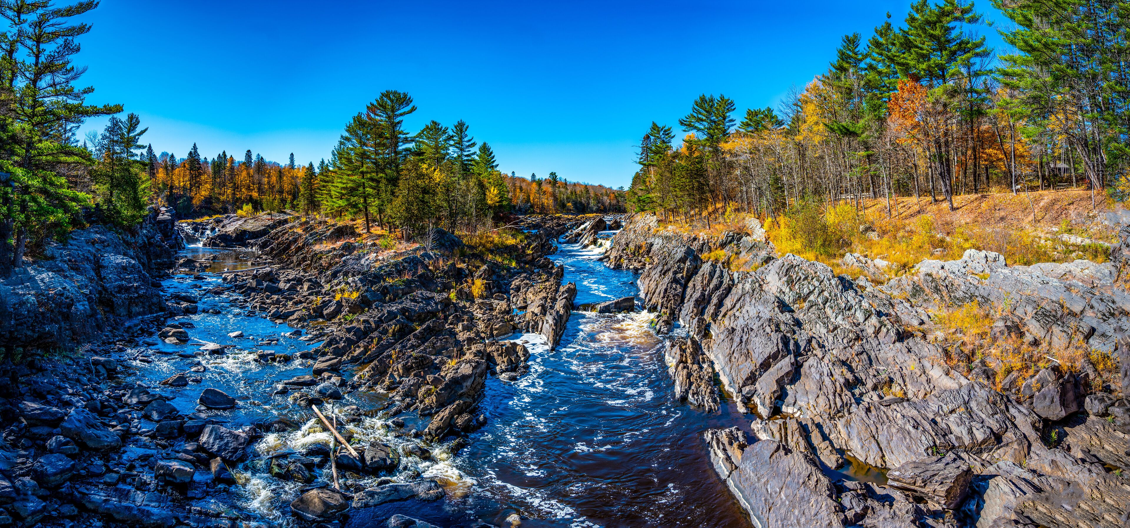 A rocky river with clear water, surrounded by dense forest and autumn-colored trees.