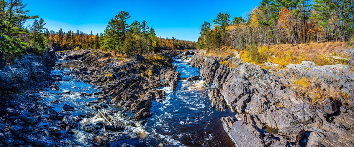 A rocky river with clear water, surrounded by dense forest and autumn-colored trees.