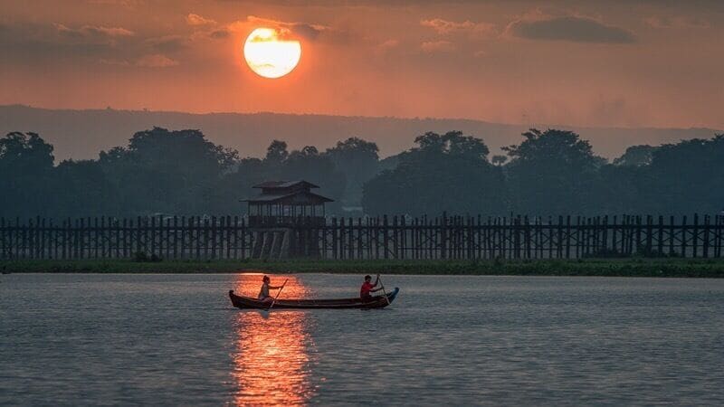 A sunset over a river with two boats, a wooden fence, and a distant structure.