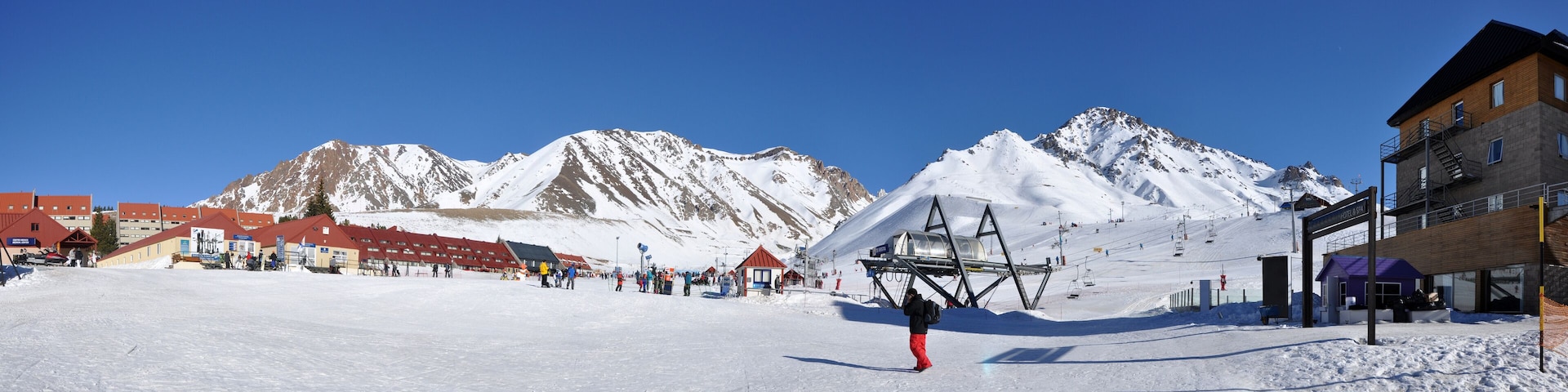 Un centro de esquí nevado con un telesilla, montañas al fondo y edificios con tejados rojos.