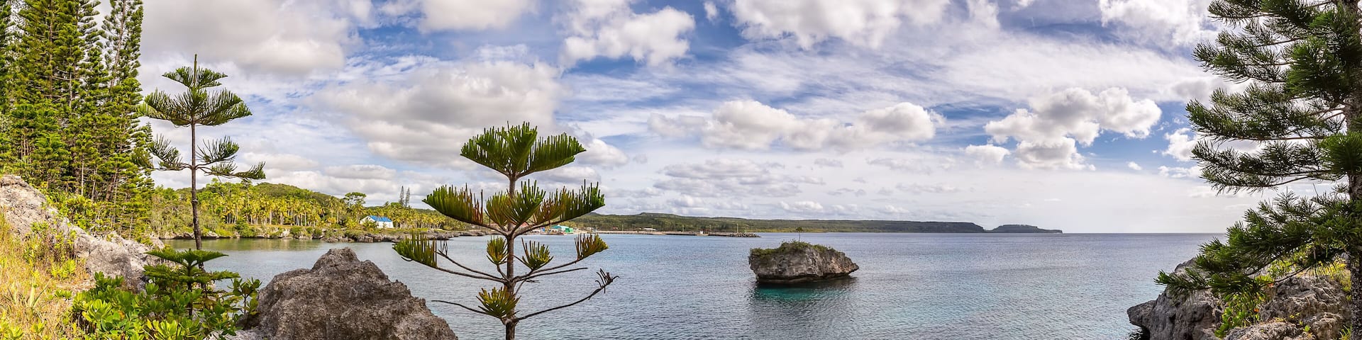 Amazing panoramic shot of Island of Mare in New Caledonia. Lone massive cliff in the water next to the island. Trees and rocks in the foreground. Blue cloudy sky as a background