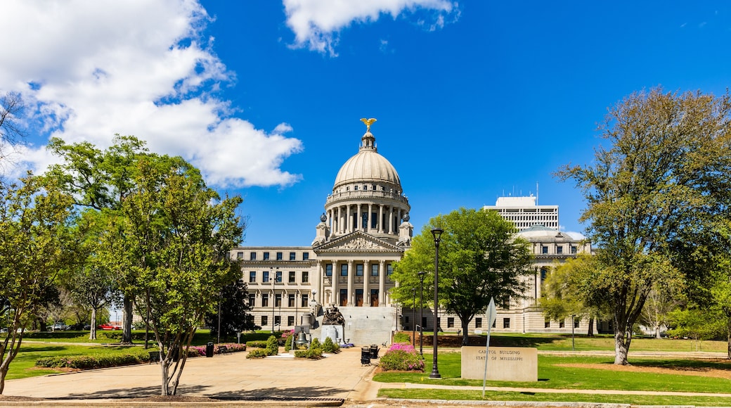 The Mississippi State Capitol building with a dome and columns, surrounded by trees and a clear blue sky.