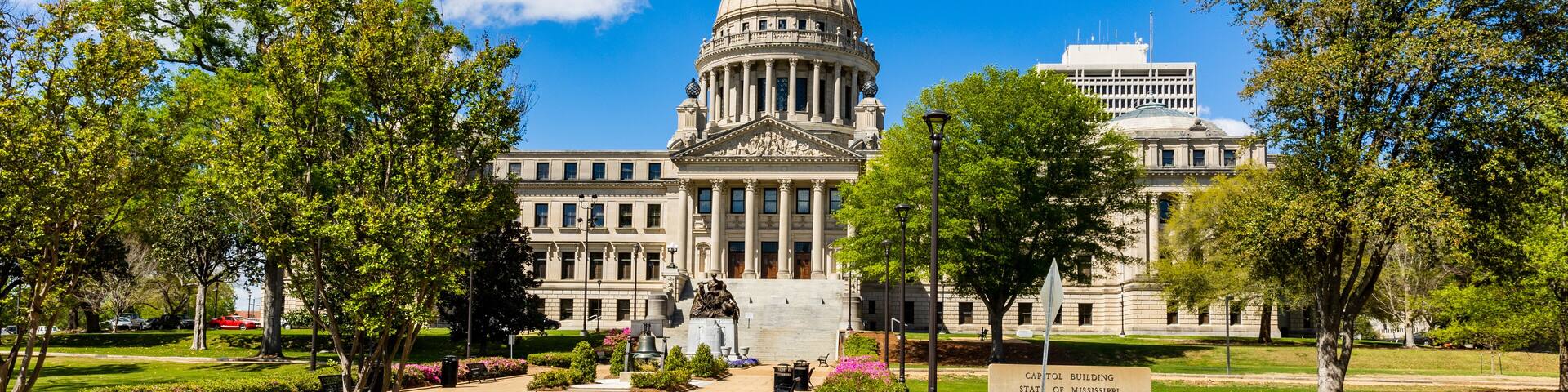 The Mississippi State Capitol building with a dome and columns, surrounded by trees and a clear blue sky.