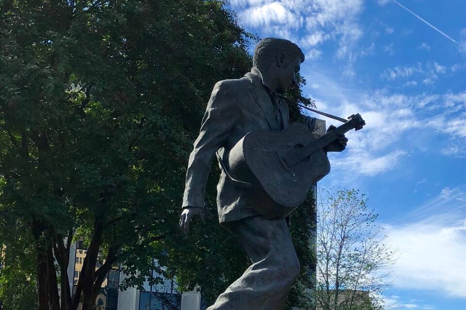 A statue of a man playing a guitar in a park with trees and a building in the background.
