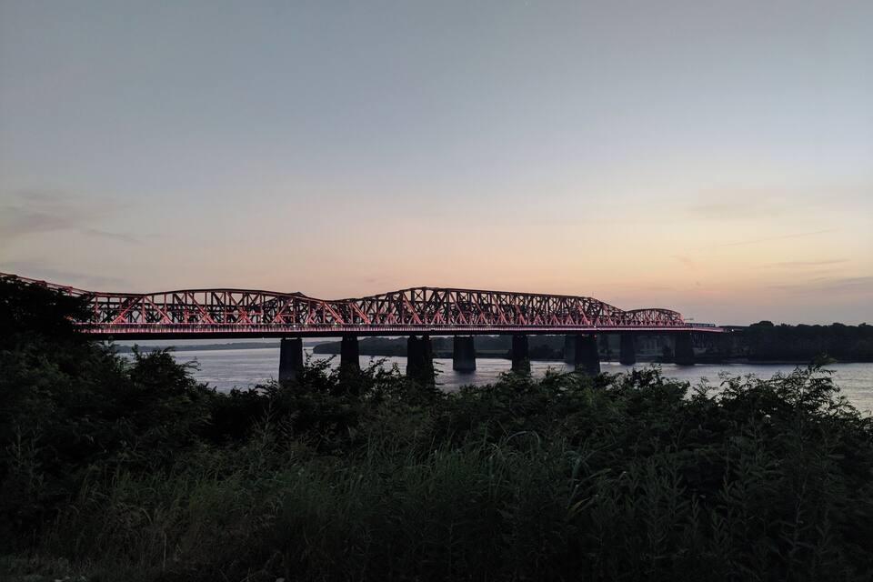 A steel bridge spanning a river with dense foliage in the foreground.