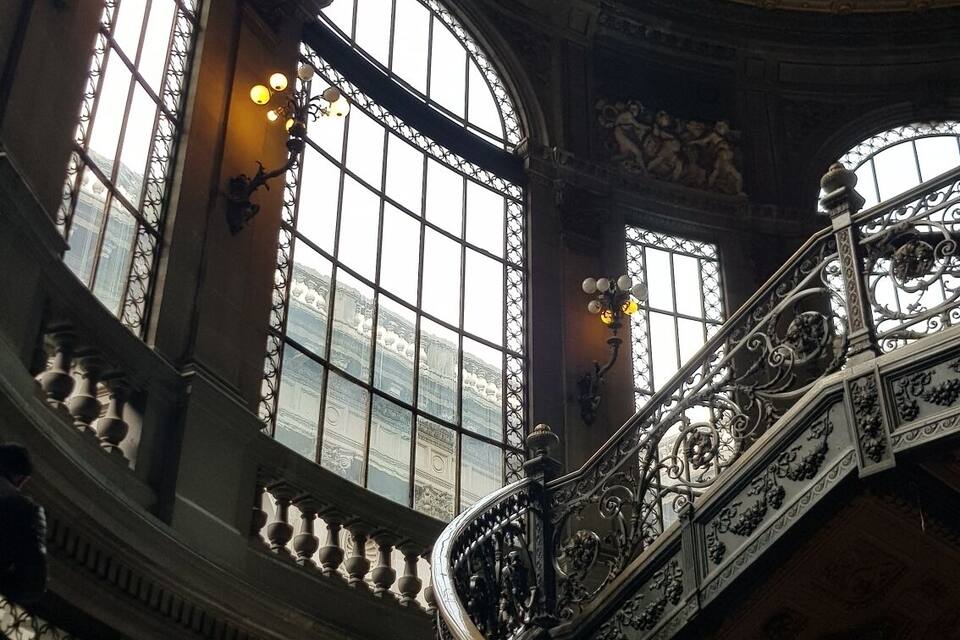 Una imponente escalera con barandales de metal ornamentados y vistas a un edificio a través de amplias ventanas arqueadas.