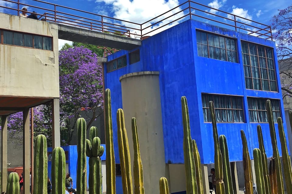 A blue building with a bridge, cacti, and purple flowers.