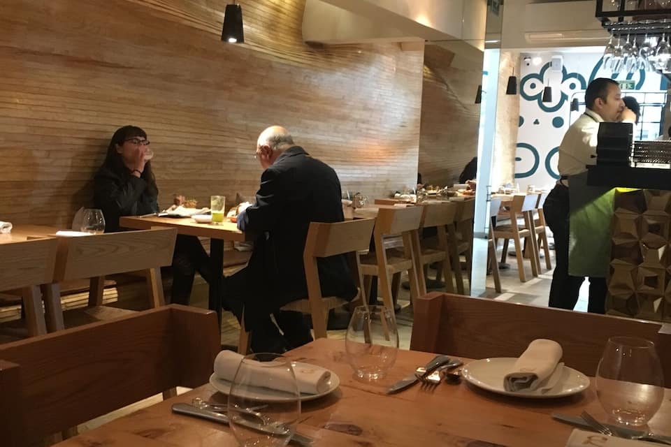A restaurant interior with wooden tables and chairs, diners, and a bar area.