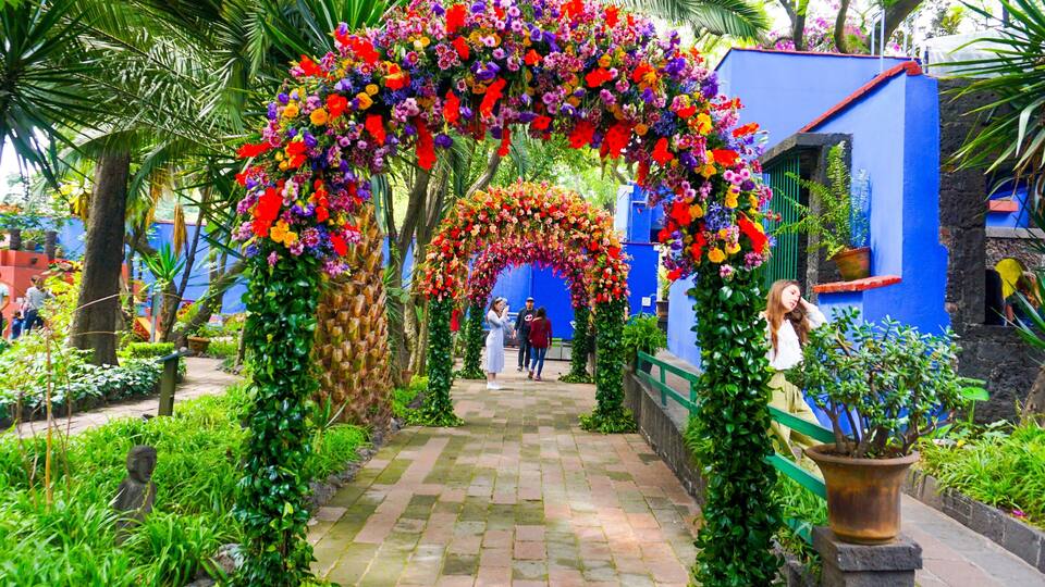 A garden with a colorful floral archway, a blue building, and a statue.