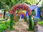 A garden with a colorful floral archway, a blue building, and a statue.