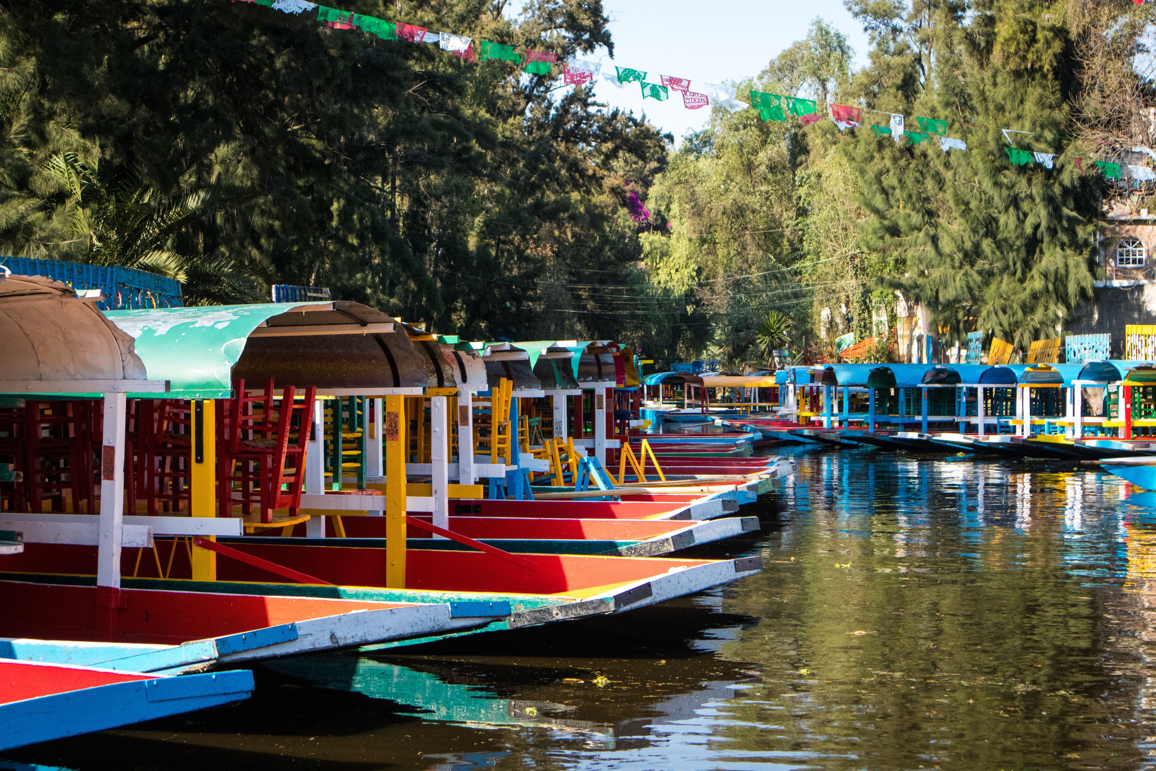 Botes coloridos amarrados a orillas de un río, rodeados de árboles y edificios.