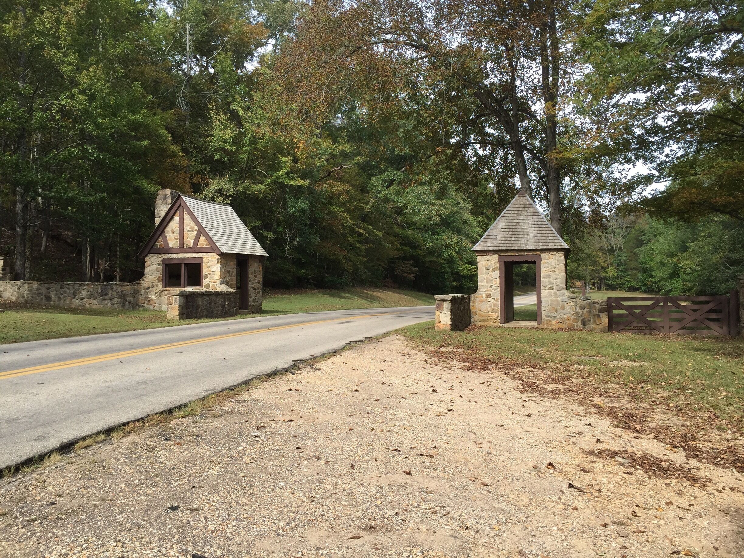 A gravel path leading to a stone structure with a triangular roof, surrounded by trees and a road.