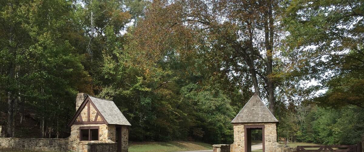 A gravel path leading to a stone structure with a triangular roof, surrounded by trees and a road.