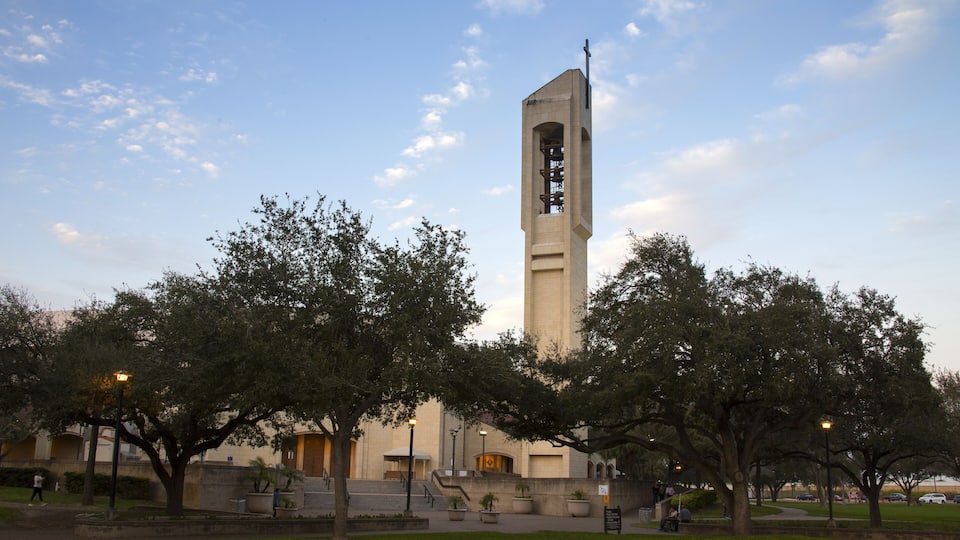 A tall tower with a cross at the top, surrounded by trees and a grassy area.
