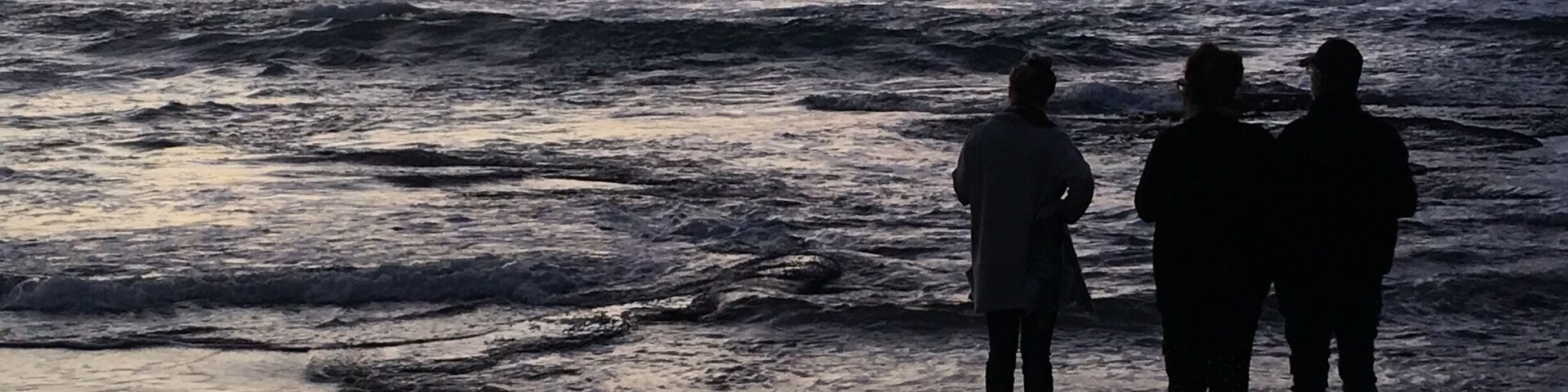 Una playa al atardecer con olas rompiendo en la orilla y tres figuras siluetas sobre la arena mojada.