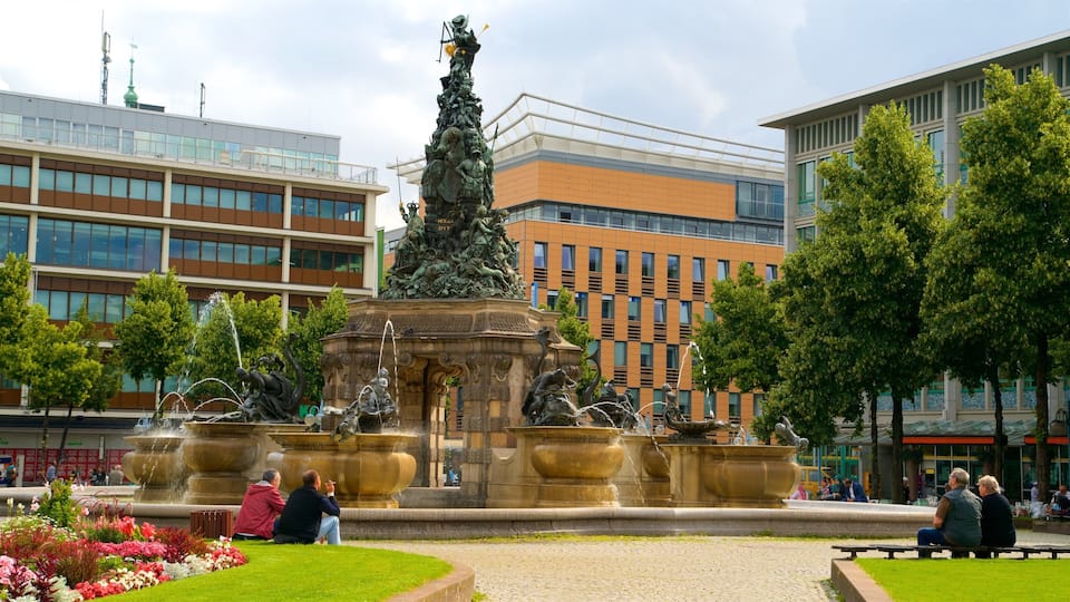 A public square with a central fountain featuring statues, surrounded by modern buildings and greenery.