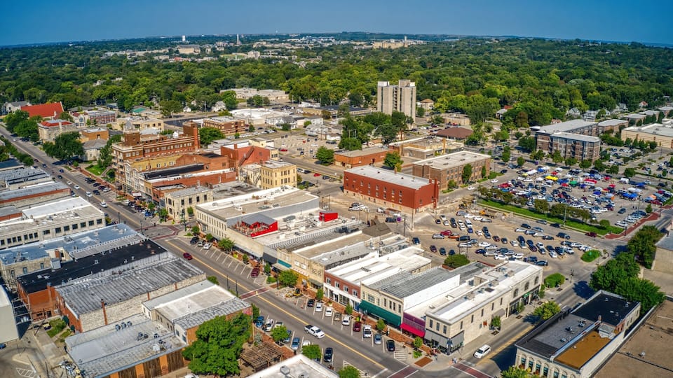 An aerial view of a historic downtown area with a mix of old and new buildings, a parking lot, and a distant view of a forested hill.