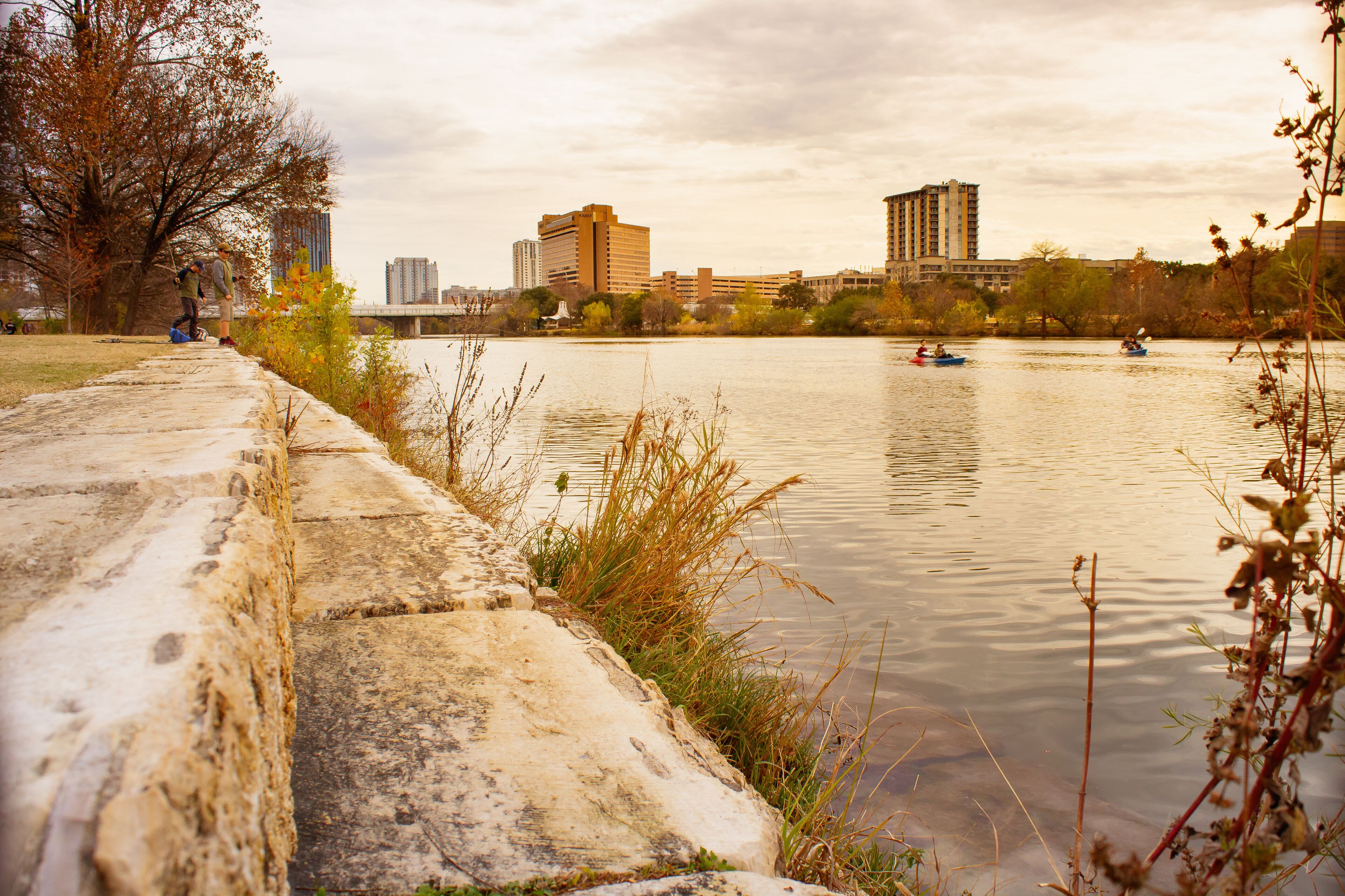 A riverside walkway with a concrete edge, a bridge in the distance, and buildings along the riverbank.
