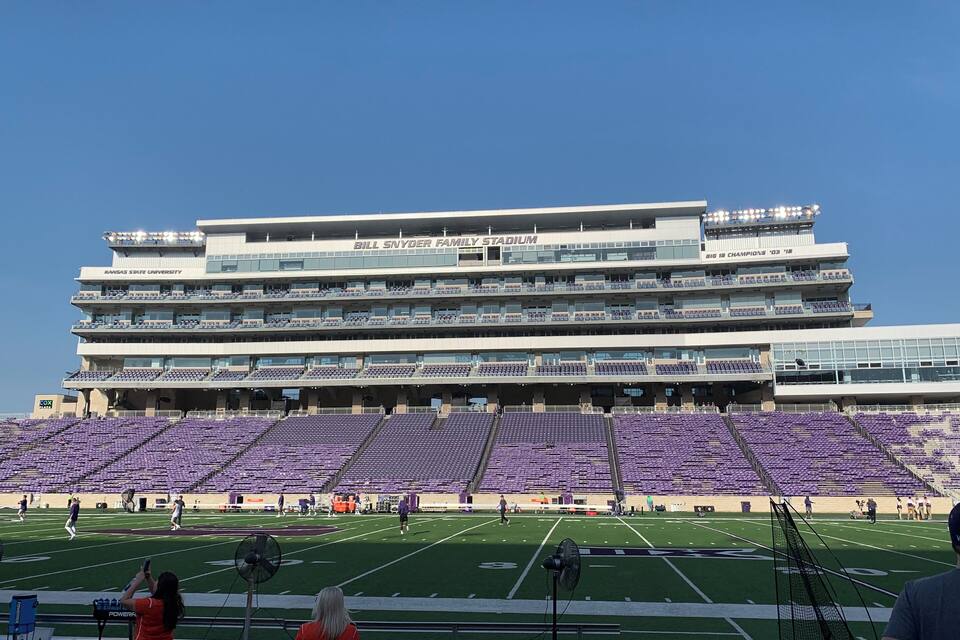 A stadium with empty purple seats, a large building with "Bill Snyder Family Stadium" and "Kansas State University" signage, and a football field.