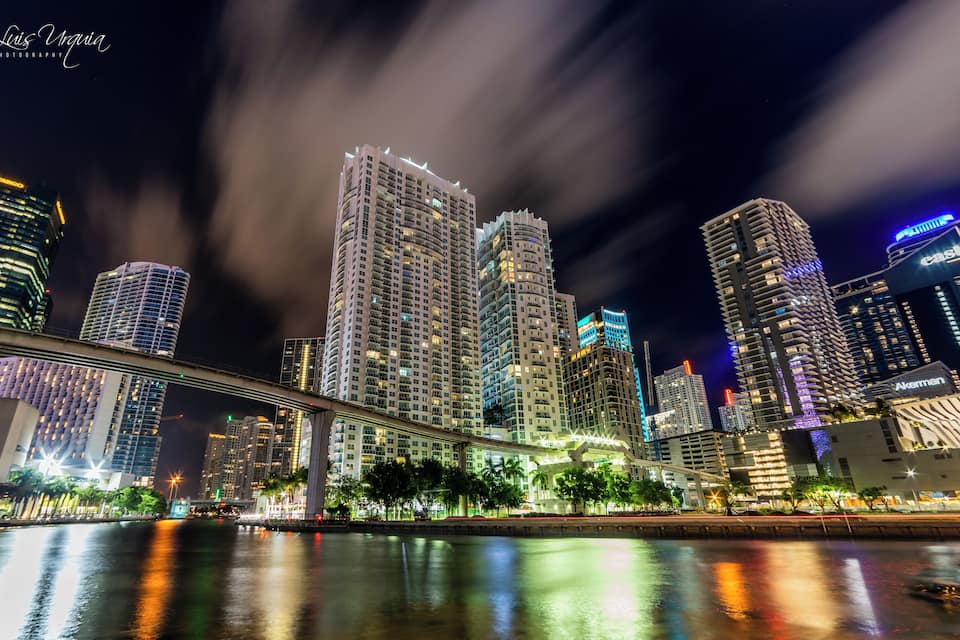 Silueta urbana nocturna amb edificis il·luminats i un pont sobre un riu.
