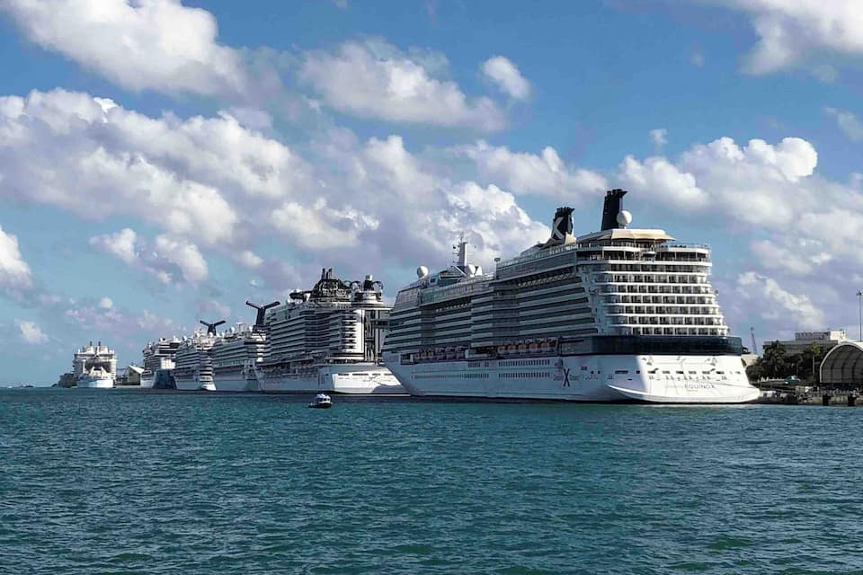 A fleet of cruise ships docked at a port with a clear sky and scattered clouds.