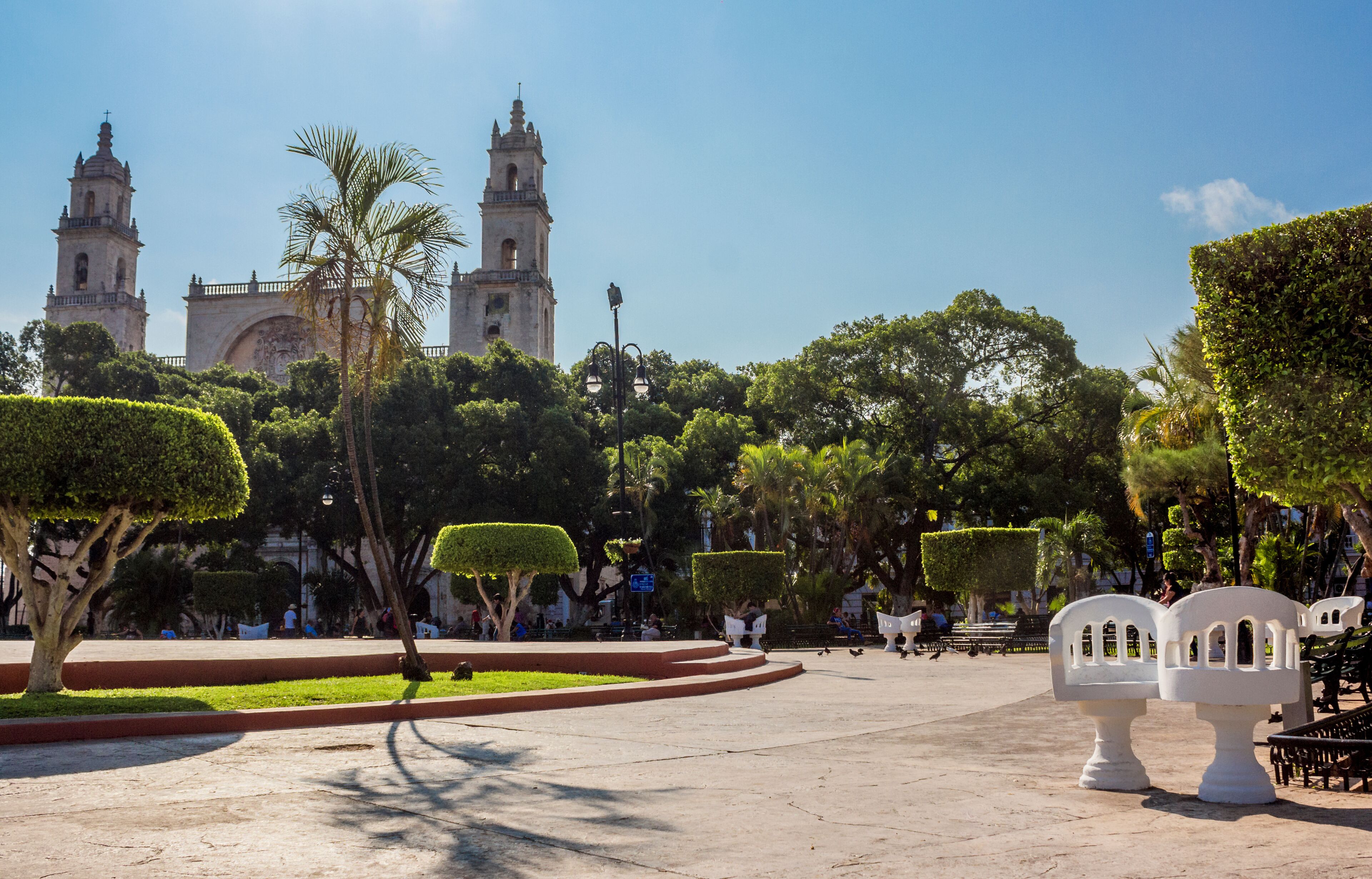 Uma praça com duas torres históricas, árvores bem podadas e bancos.