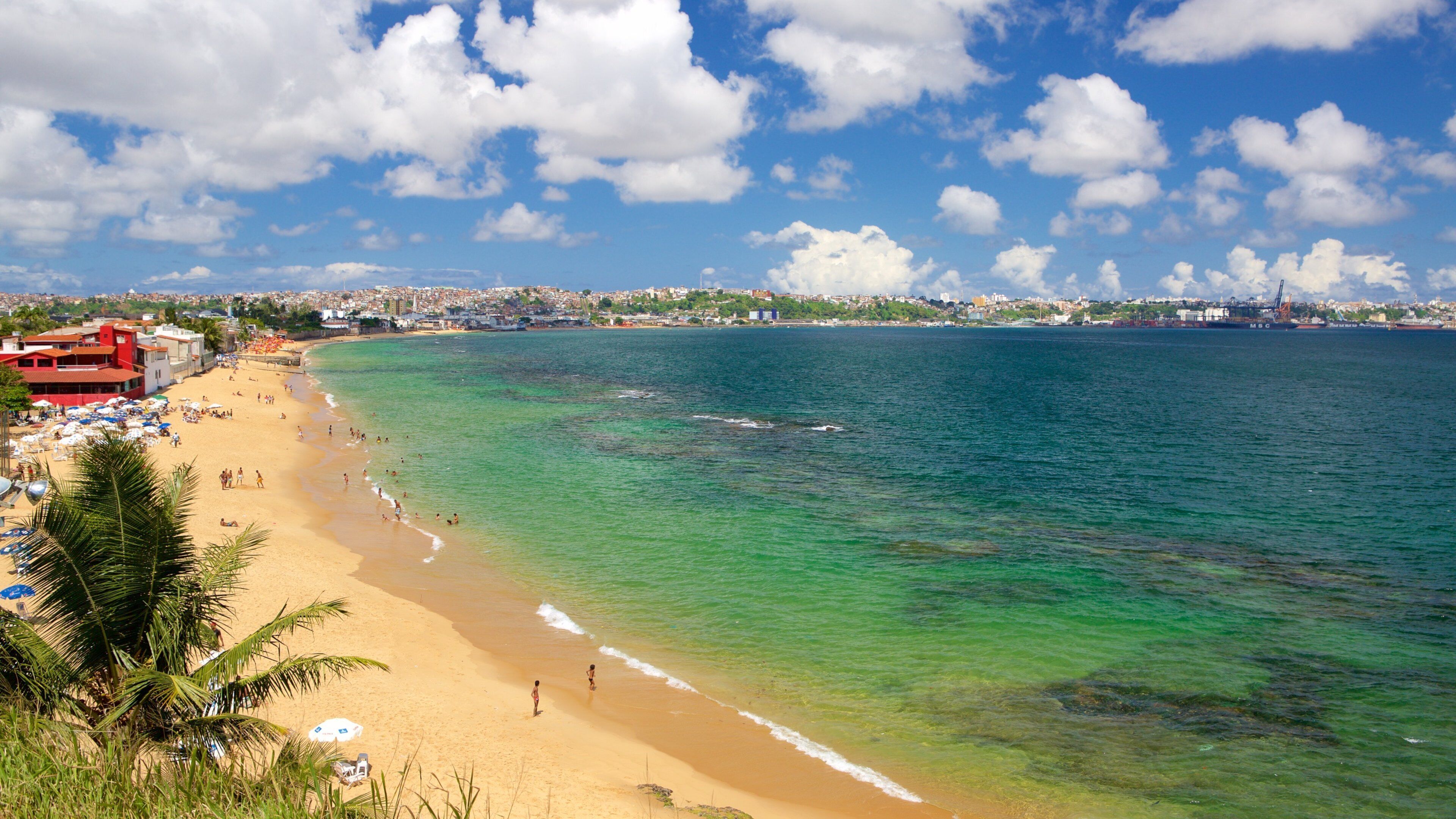 A beach with clear turquoise waters, a sandy shore, and a cityscape in the background.