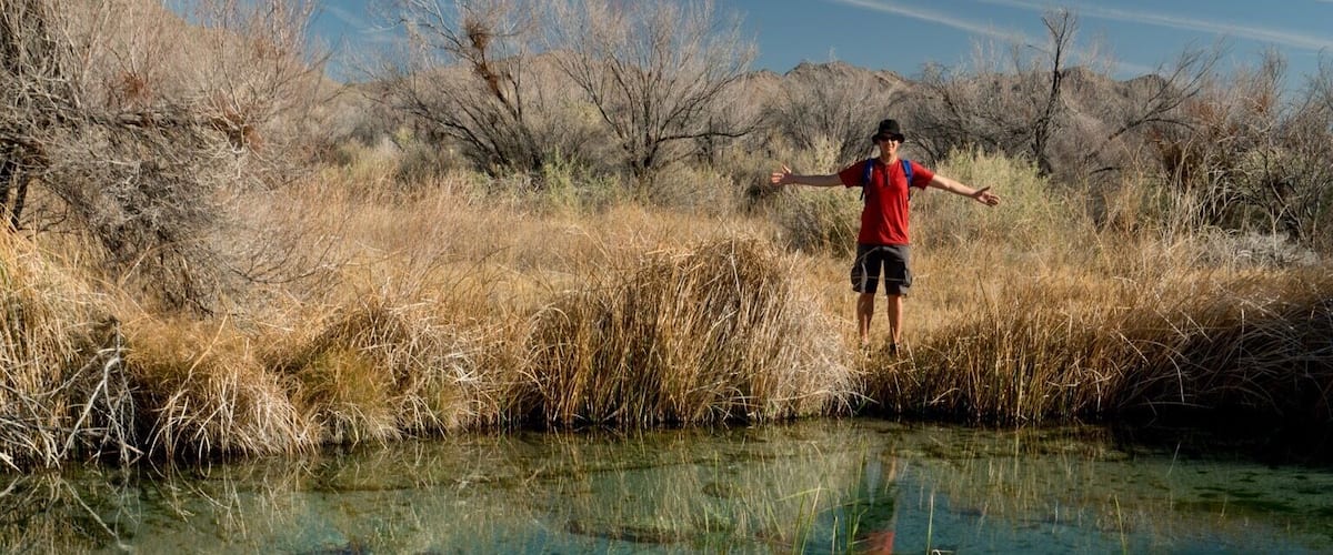 A person standing in a clear shallow water body surrounded by dry grass and bushes.
