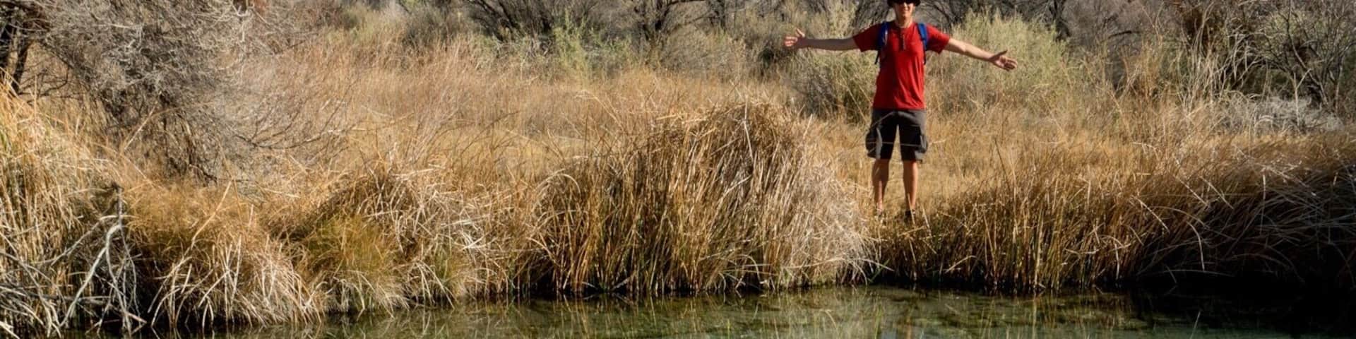 A person standing in a clear shallow water body surrounded by dry grass and bushes.