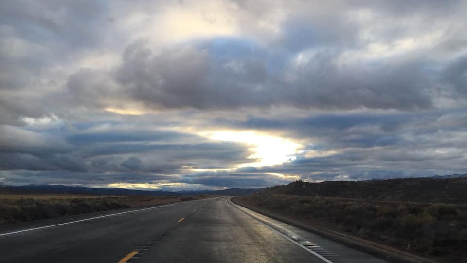 A road curving through a desert landscape with a cloudy sky and the sun peeking through the clouds.