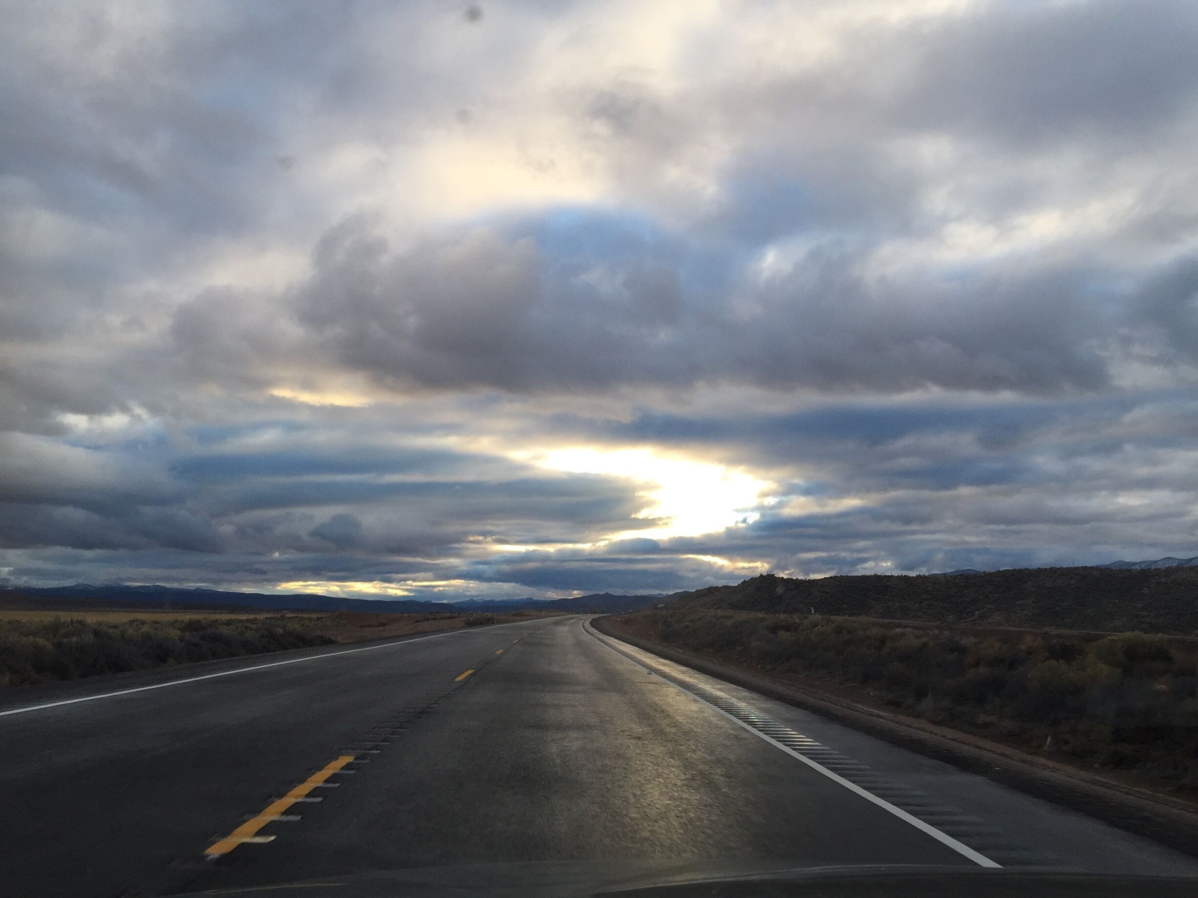 A road curving through a desert landscape with a cloudy sky and the sun peeking through the clouds.