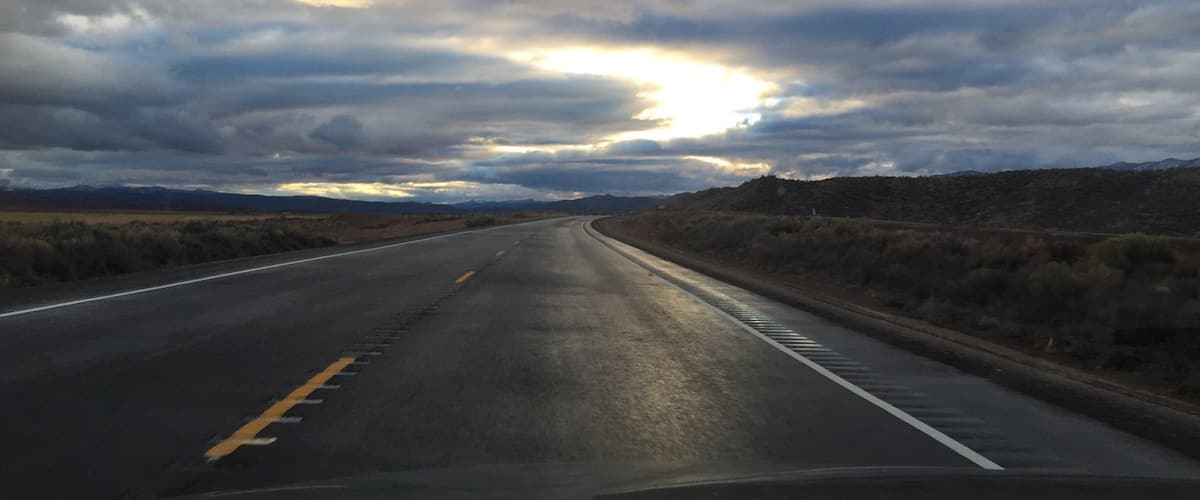 A road curving through a desert landscape with a cloudy sky and the sun peeking through the clouds.