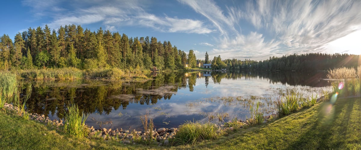 A serene lake surrounded by dense forest, with a house on the opposite shore and a clear sky with wispy clouds.
