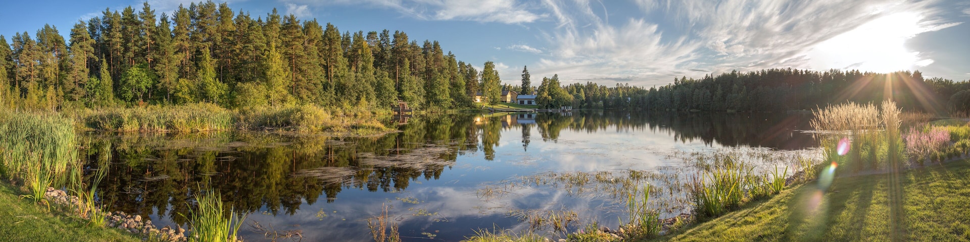 A serene lake surrounded by dense forest, with a house on the opposite shore and a clear sky with wispy clouds.