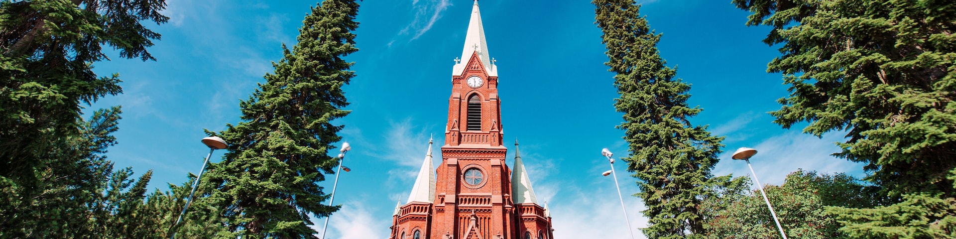Una iglesia de ladrillo rojo con un campanario, rodeada de altos árboles de hoja perenne y un cielo azul despejado.