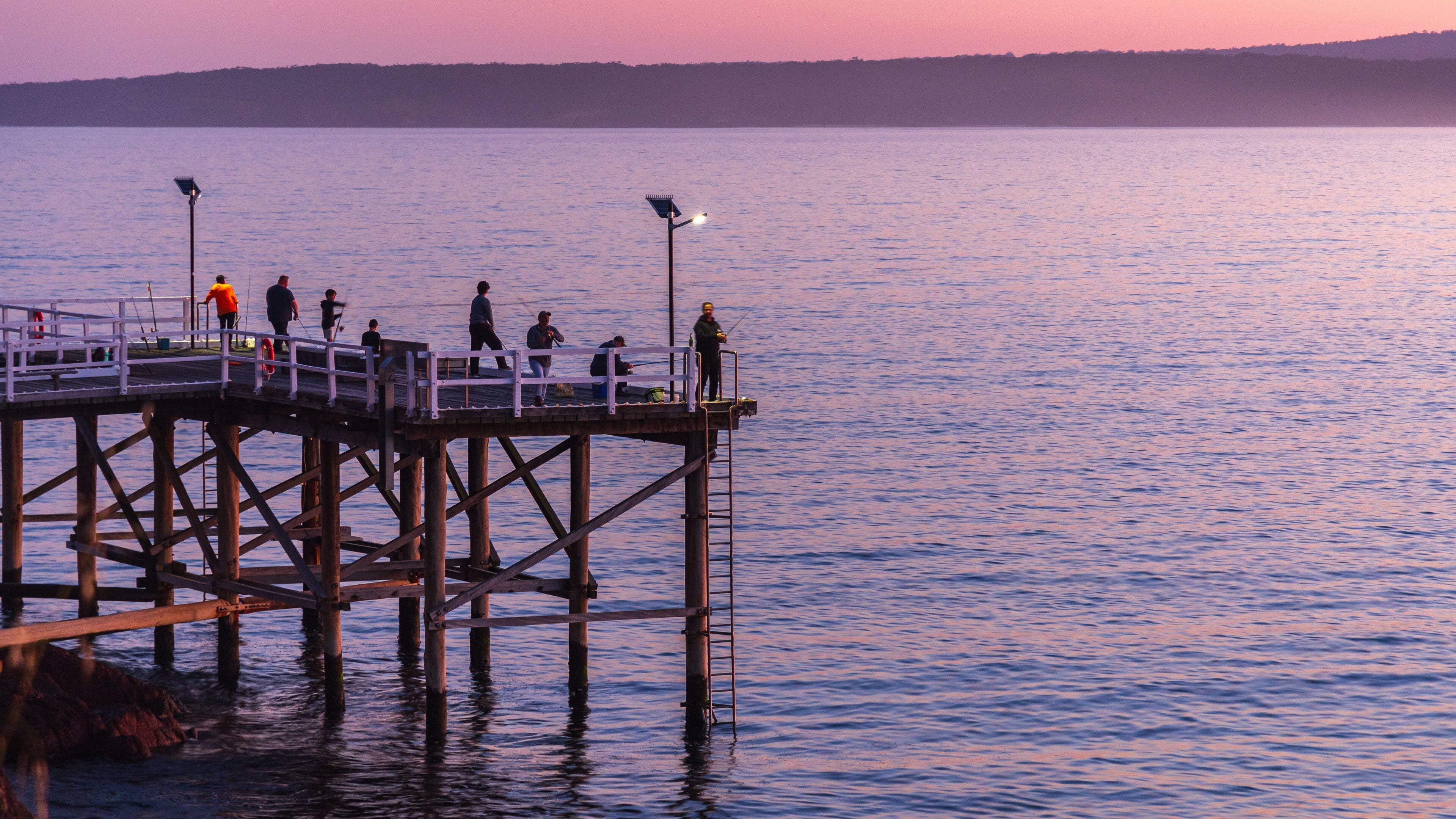 A pier extending into calm water at sunset with people fishing and walking.