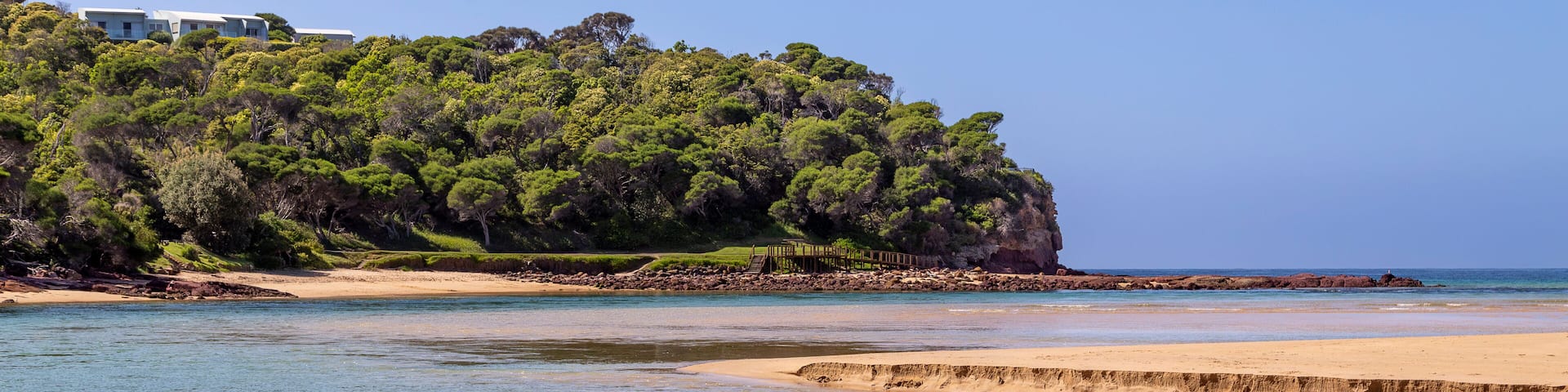 A coastal landscape with a sandy beach, calm waters, and a forested hill.